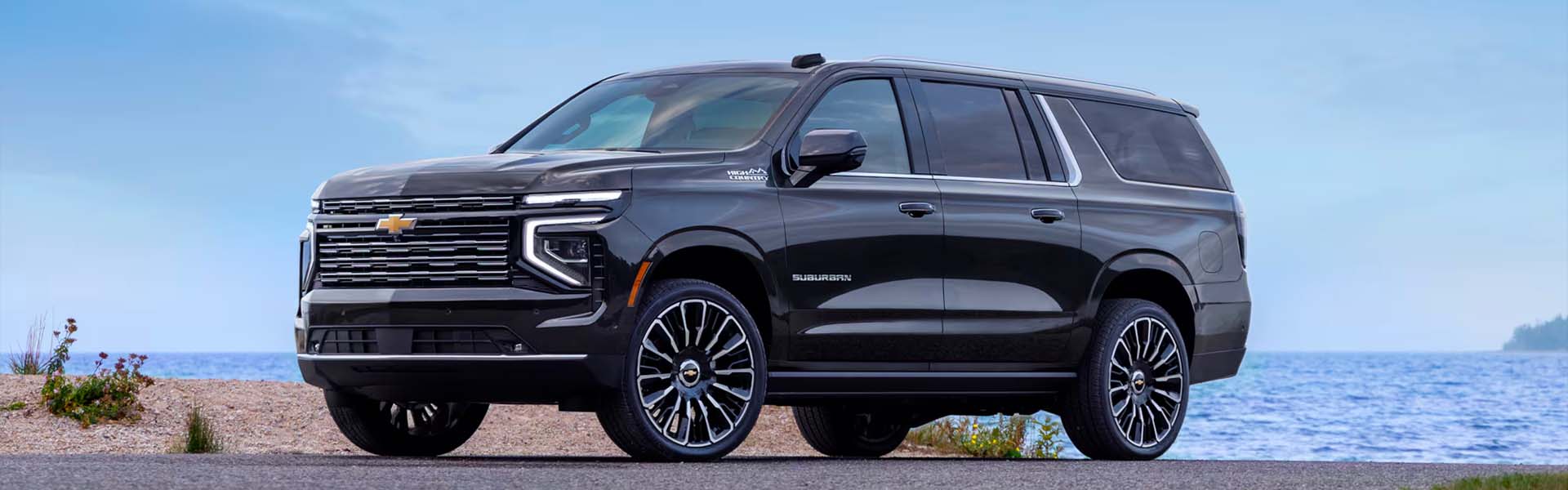2025 Chevrolet Suburban parked near a waterfront with a clear sky, showcasing its bold front grille, black exterior, and premium SUV design.