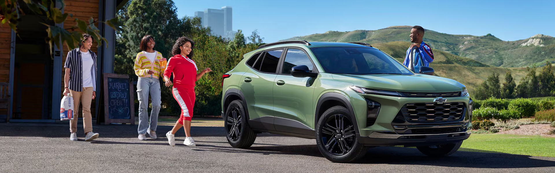 2026 Chevrolet Trax compact SUV in green parked near a roadside market with people walking and mountains in the distance.
