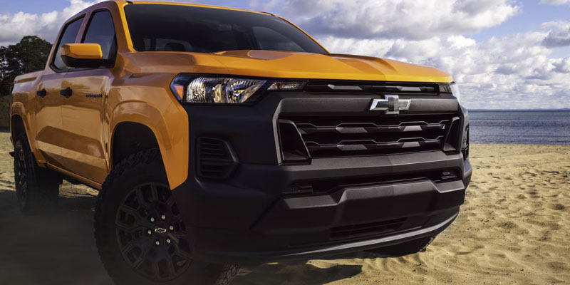A bright yellow pickup truck Colorado is parked on a sandy beach under a partly cloudy sky. The truck's bold grille and rugged design convey strength.