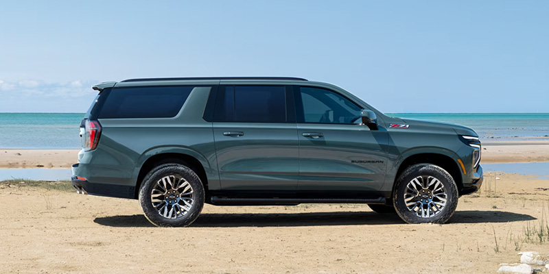 A dark green Suburban is parked on a sandy beach, facing right. The ocean and blue sky form a serene backdrop, emphasizing a sense of adventure and calm.